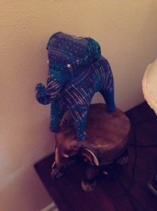 Stuffed fabric elephant statue with cross hatched blue, black, and grey stripes. Elephant is standing on a carved wood elephant plant stand.