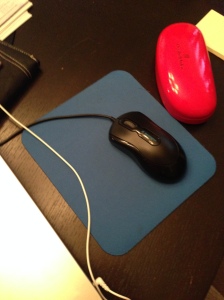 Blue mousepad on a black table. A black mouse sits atop the mousepad, and a red glasses case sits to the right.