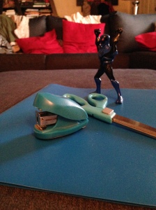 Blue folder underneath a blue mini stapler, blue handled scissors, and a blue and black Venom action figure.